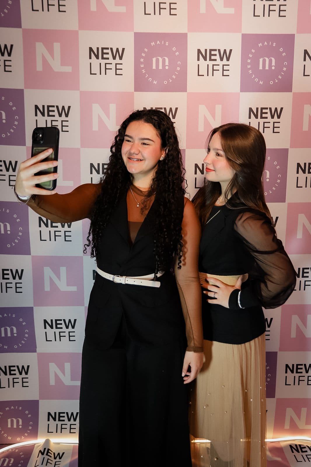 Duas jovens tirando uma selfie diante do painel da New Life, no espaço de fotos do evento.