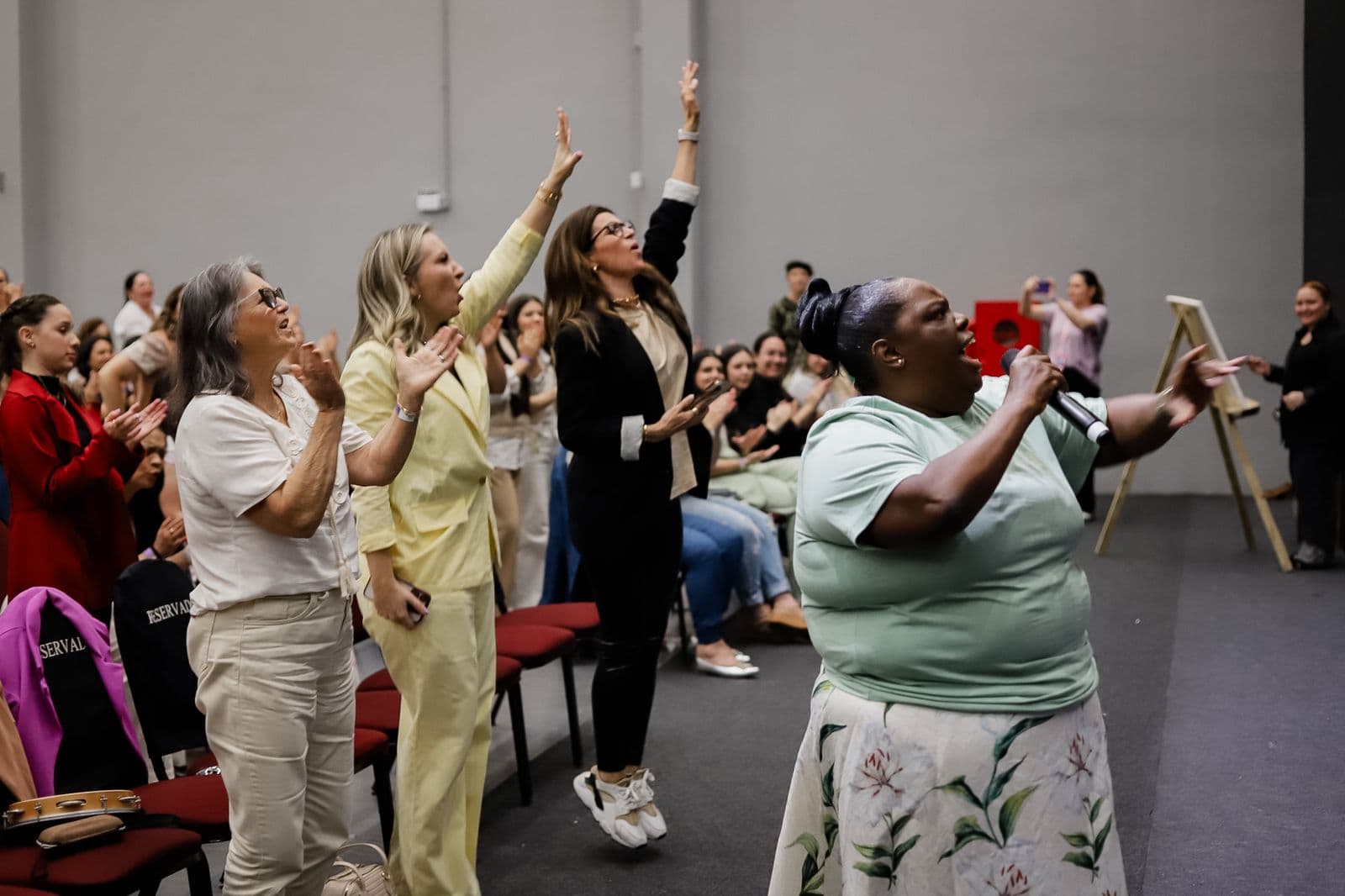 Mulheres em pé no meio do público, cantando e com as mãos erguidas em um momento de adoração.
