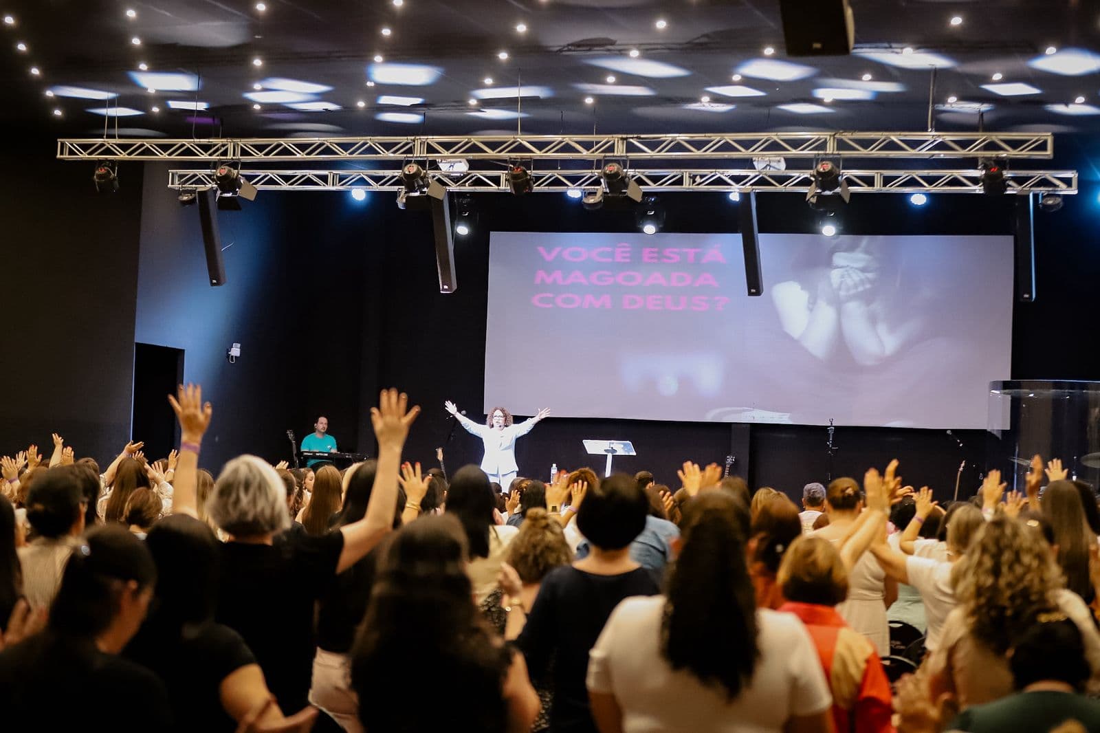 Auditório lotado de mulheres com as mãos levantadas em adoração, com a preletora no palco e o telão ao fundo.